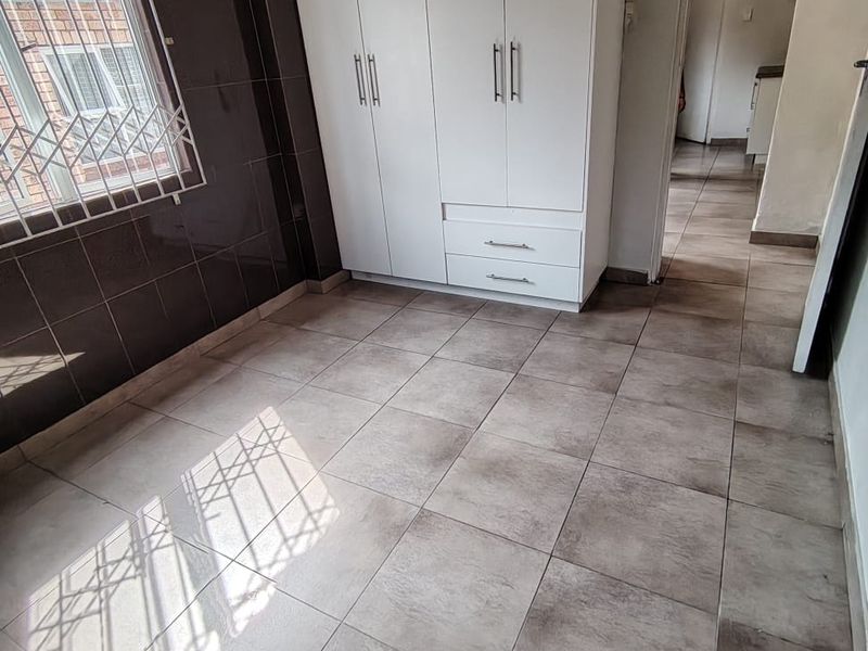 A bright, empty bedroom featuring neutral floor tiles, a built-in white wardrobe, and a window fitted with security bars.