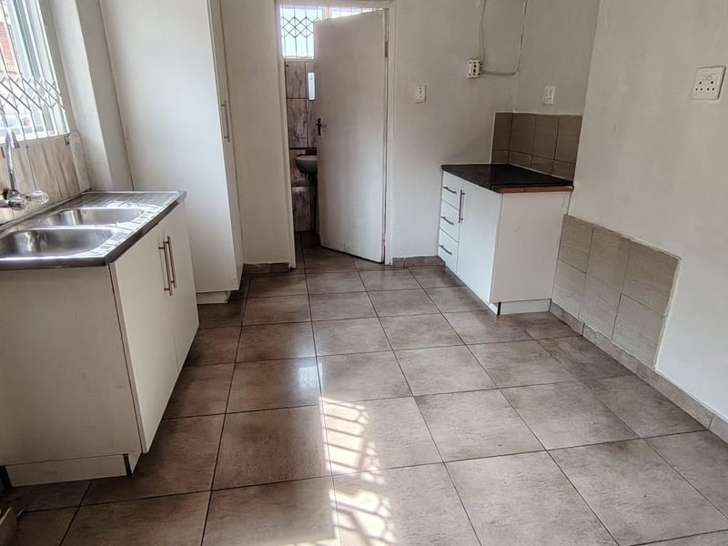 A functional, empty kitchen featuring tiled flooring, built-in cabinetry, and a double stainless steel sink, with direct access to an adjacent room.