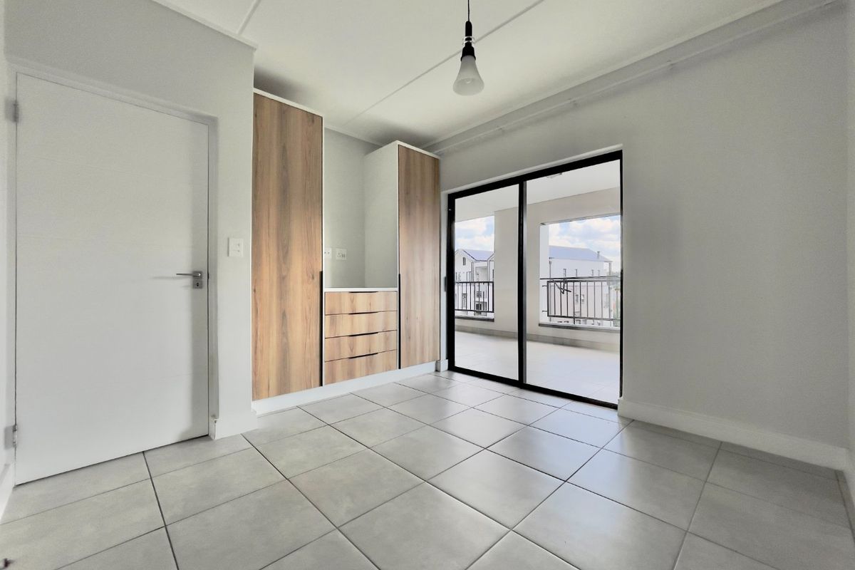 A bright, empty bedroom featuring modern built-in cupboards, neutral tiled flooring, and a sliding glass door leading to a private balcony.