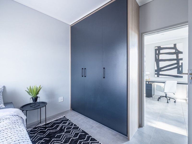 A modern bedroom featuring a large, dark-toned built-in wardrobe and neutral tiled flooring, with a view into an adjacent office space.