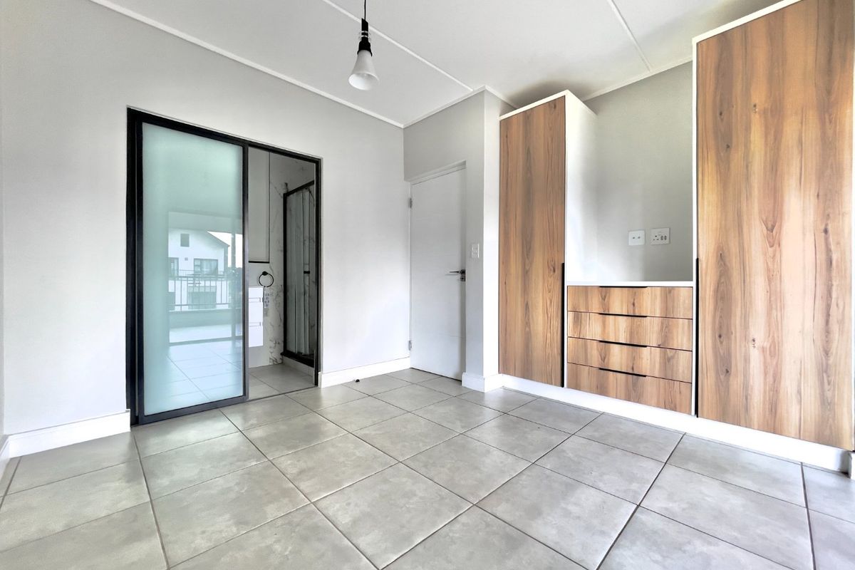 A bright, modern bedroom featuring neutral tiled flooring, built-in wooden cabinetry, and a sliding glass door leading to an en-suite bathroom.