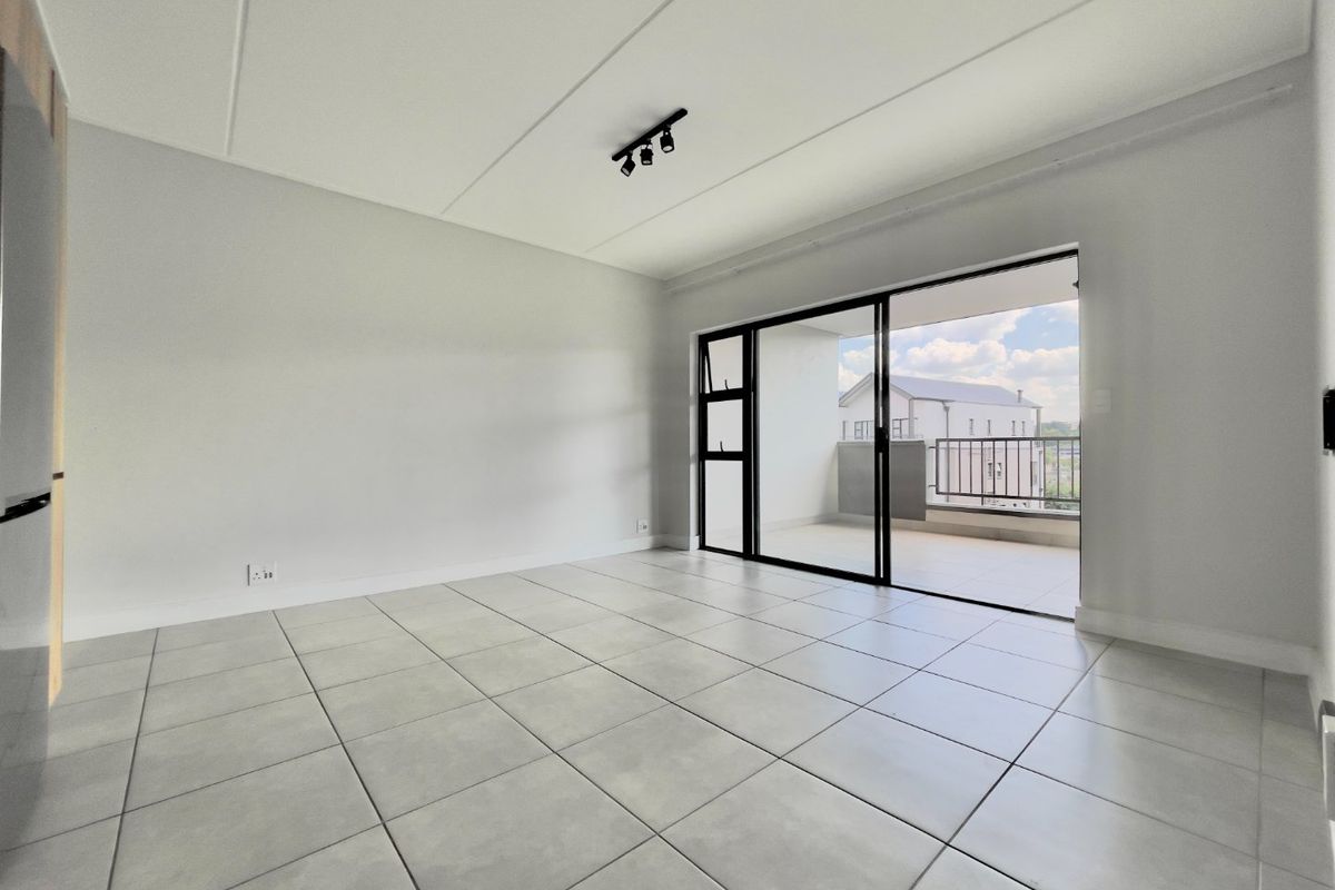 A bright, empty living area featuring neutral tiled flooring, modern track lighting, and large sliding glass doors that open onto a private balcony.