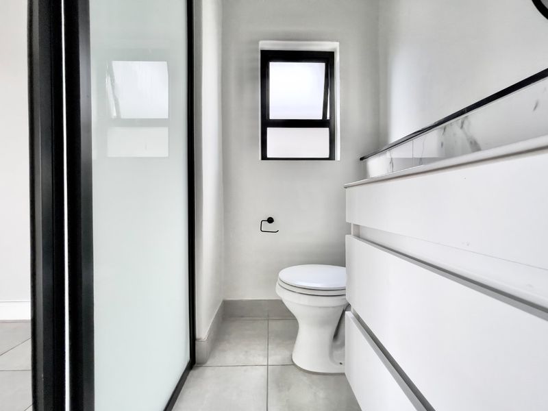A modern, minimalist bathroom featuring a sleek white vanity with marble-look countertop, a toilet, and a contemporary black-framed shower screen.
