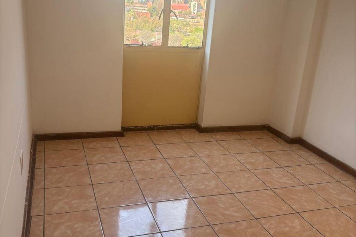 An empty bedroom featuring ceramic floor tiles, skirting boards, and an arched window with a view of the surrounding urban area.