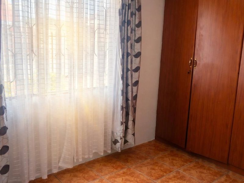 A bright, empty bedroom featuring built-in wooden wardrobes, ceramic tiled flooring, and a window fitted with security bars.