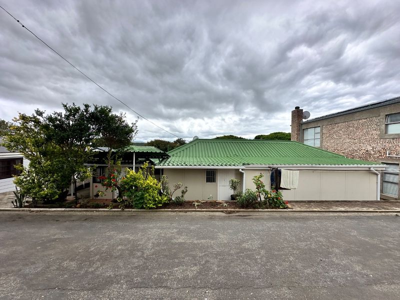 Front elevation of a single-story house featuring a green corrugated metal roof, brick chimney, and paved driveway access.