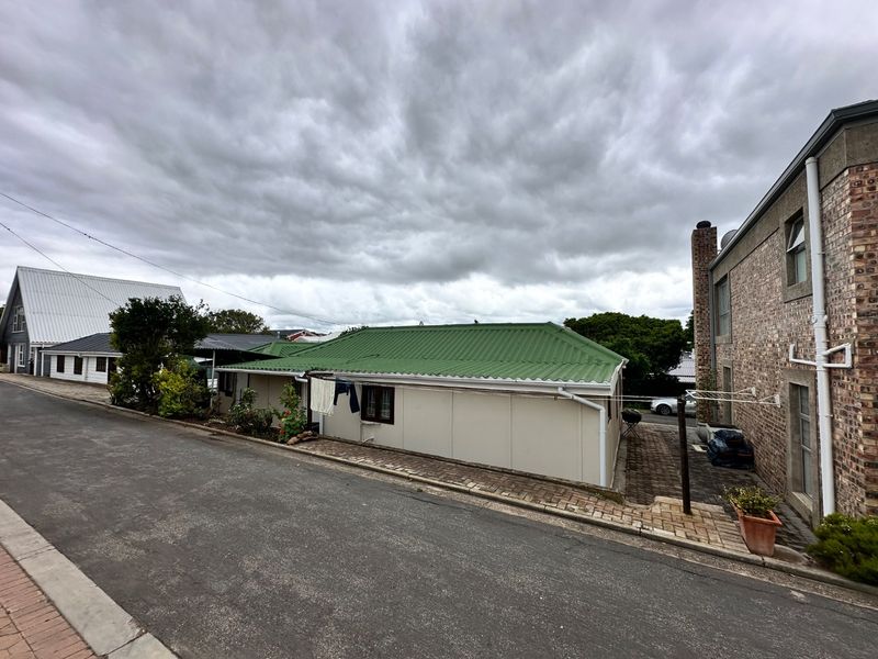 A street-level view of the property featuring a green corrugated metal roof, face brick exterior, and paved driveway area under a cloudy sky.