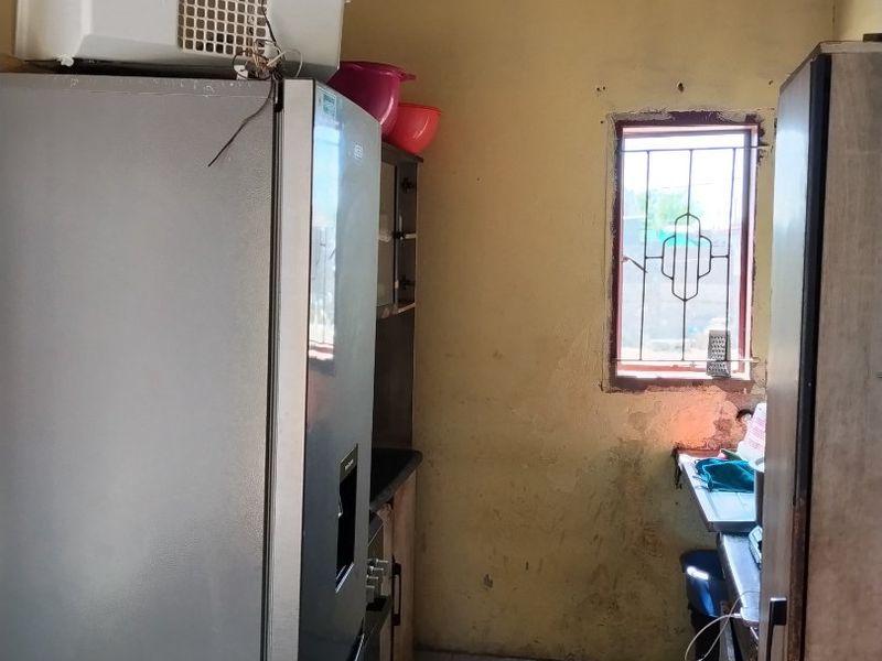 A small kitchen area requiring significant renovation, featuring tiled floors, a window with security bars, and dated cabinetry with visible wall and ceiling deterioration.