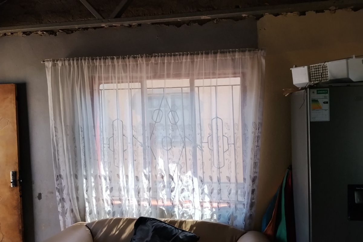 A living area featuring a window with security bars and exposed ceiling structure, requiring finishing work on walls and ceiling.