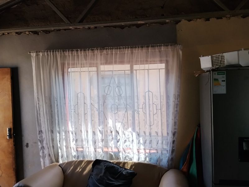 A living area featuring a window with security bars and exposed ceiling structure, requiring finishing work on walls and ceiling.