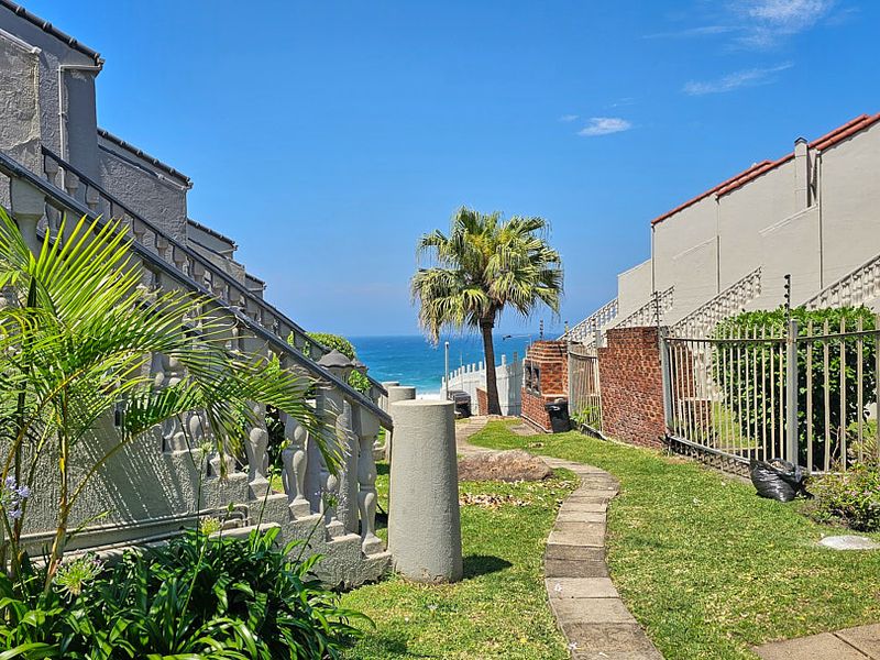 A rear exterior view of the apartment complex featuring a paved walkway, stone balustrades, and a direct path leading toward the ocean.