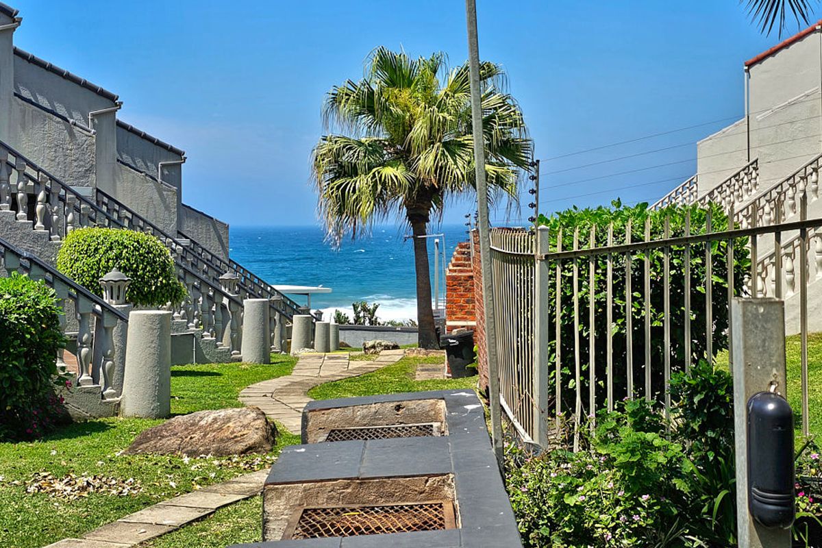 A well-maintained rear garden area featuring a built-in braai, paved pathway, and concrete balustrades, offering a scenic view of the ocean.