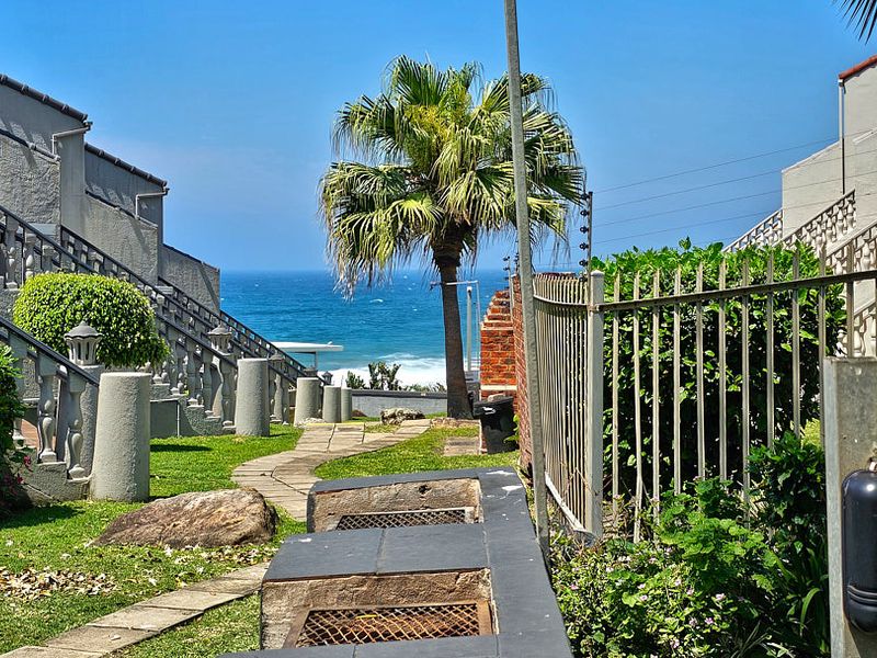 A well-maintained rear garden area featuring a built-in braai, paved pathway, and concrete balustrades, offering a scenic view of the ocean.