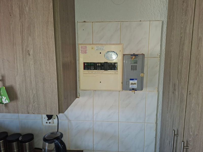 A kitchen wall section featuring a built-in electrical distribution board and a prepaid electricity meter mounted on tiled splashback between laminate kitchen cabinets.