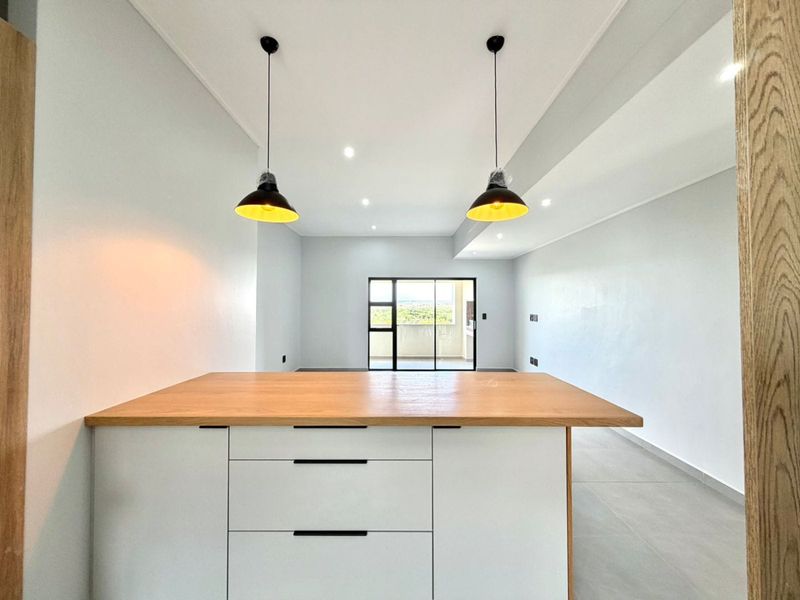 Modern open-plan living area featuring a kitchen island with a wooden countertop, pendant lighting, and neutral tiled flooring leading to a balcony.