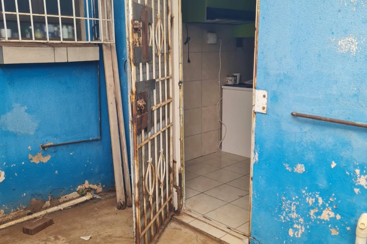 Rear exterior view showing a weathered blue wall, a rusted security gate, and a glimpse into a tiled interior space.