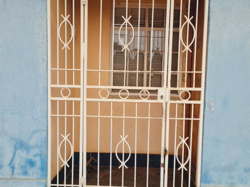 Exterior entrance featuring a white metal security gate, tiled threshold, and blue painted wall with visible signs of wear and weathering.