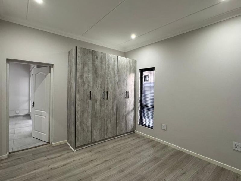 A bright, modern bedroom featuring light wood-look laminate flooring, neutral walls, and contemporary built-in cupboards with a textured grey finish.