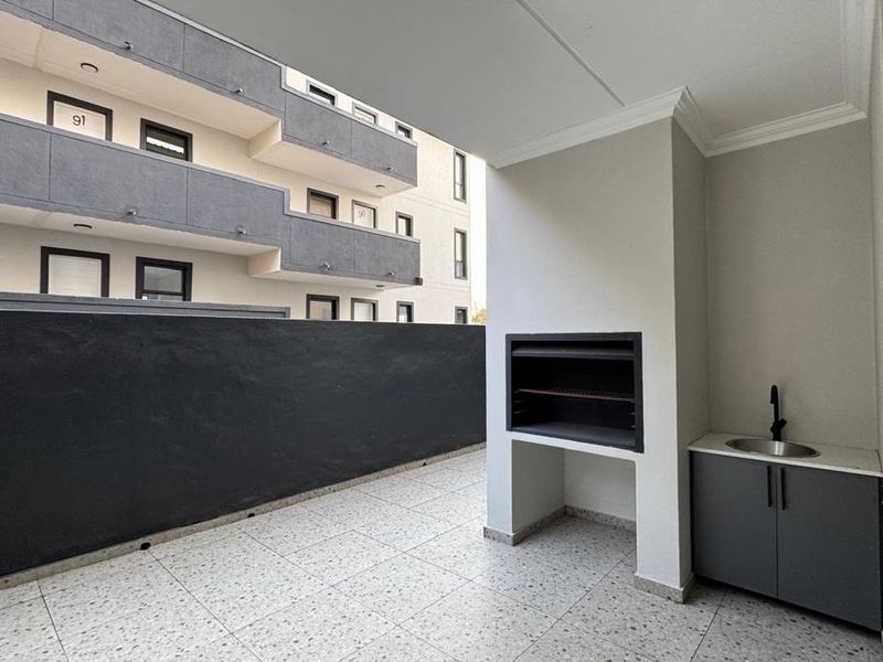 A modern covered balcony featuring a built-in braai, a small sink with cabinetry, and clean tiled flooring, offering a functional outdoor entertainment space.