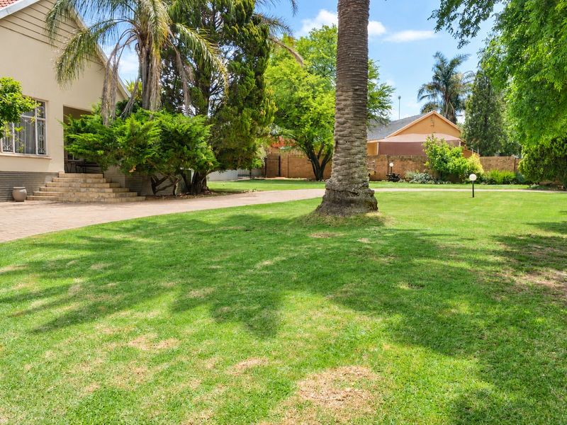 A spacious, well-maintained garden featuring a large lawn, mature trees, a paved driveway, and a brick boundary wall.