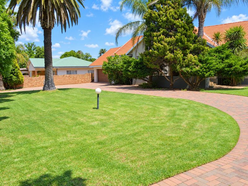 A well-maintained front garden featuring a manicured lawn, mature palm trees, and a paved driveway leading to the garage.