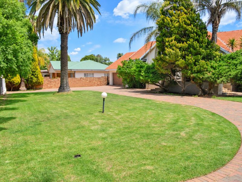 A well-maintained front garden featuring a brick-paved driveway, established palm trees, and a neat lawn area leading to the main house entrance.