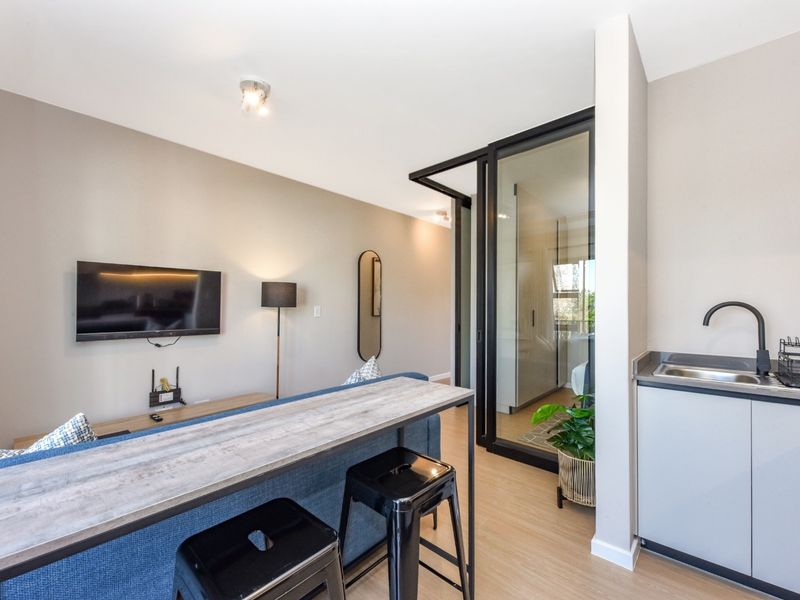 Modern open-plan living area featuring a compact kitchenette with sink, laminate flooring, and a sleek glass partition door leading to the bedroom.