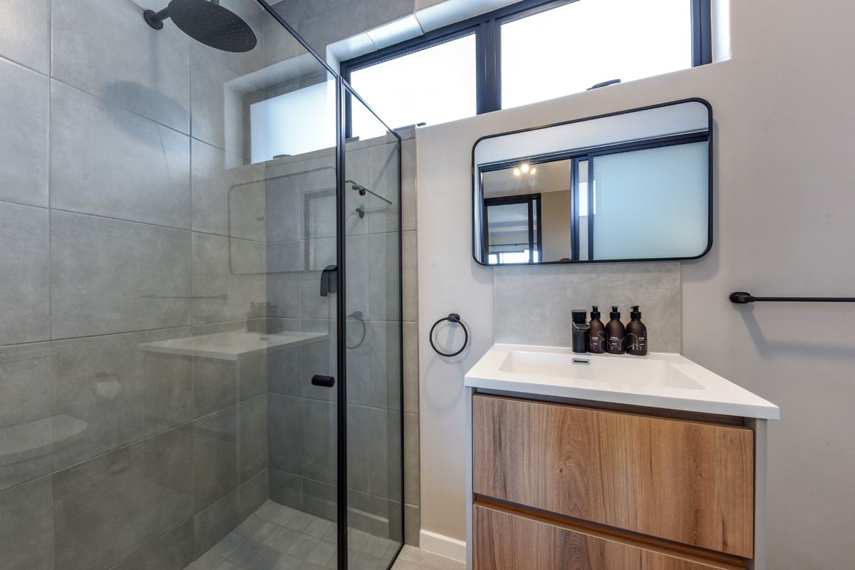 Modern bathroom featuring a walk-in shower with black fixtures, a wood-finish vanity cabinet, and a sleek wall-mounted mirror under clerestory windows.