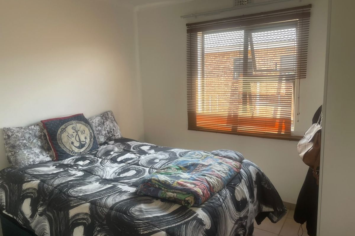A functional bedroom featuring tiled flooring, a window with fitted venetian blinds, and a wall-mounted ventilation grille.