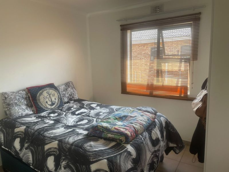 A functional bedroom featuring tiled flooring, a window with fitted venetian blinds, and a wall-mounted ventilation grille.