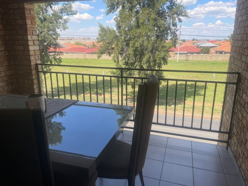 A covered balcony featuring brick pillars, tiled flooring, and a metal balustrade, offering an open view of the surrounding neighborhood.