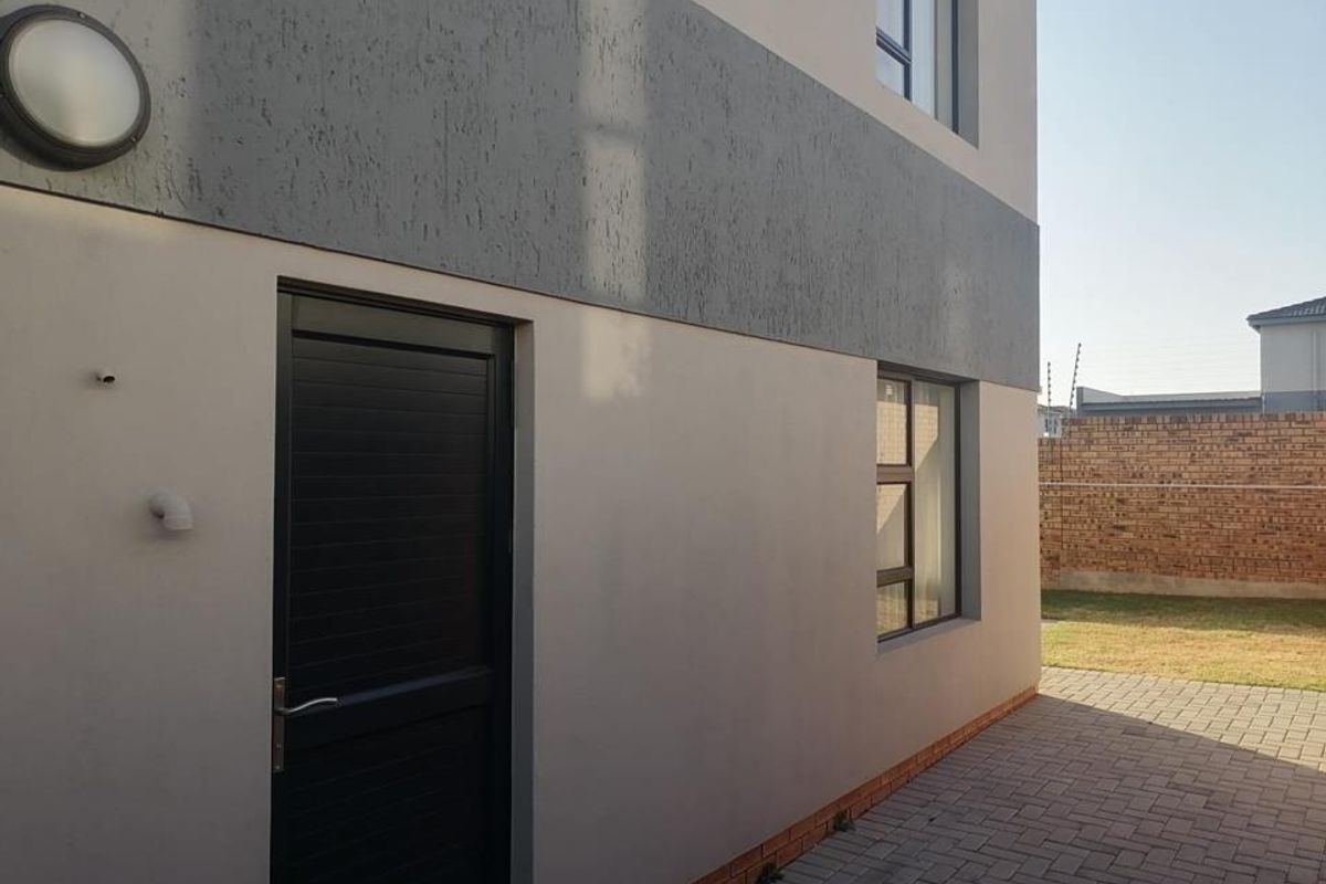 A clean rear exterior view featuring a paved patio area, modern plastered walls with contrasting grey detailing, and aluminium window frames.