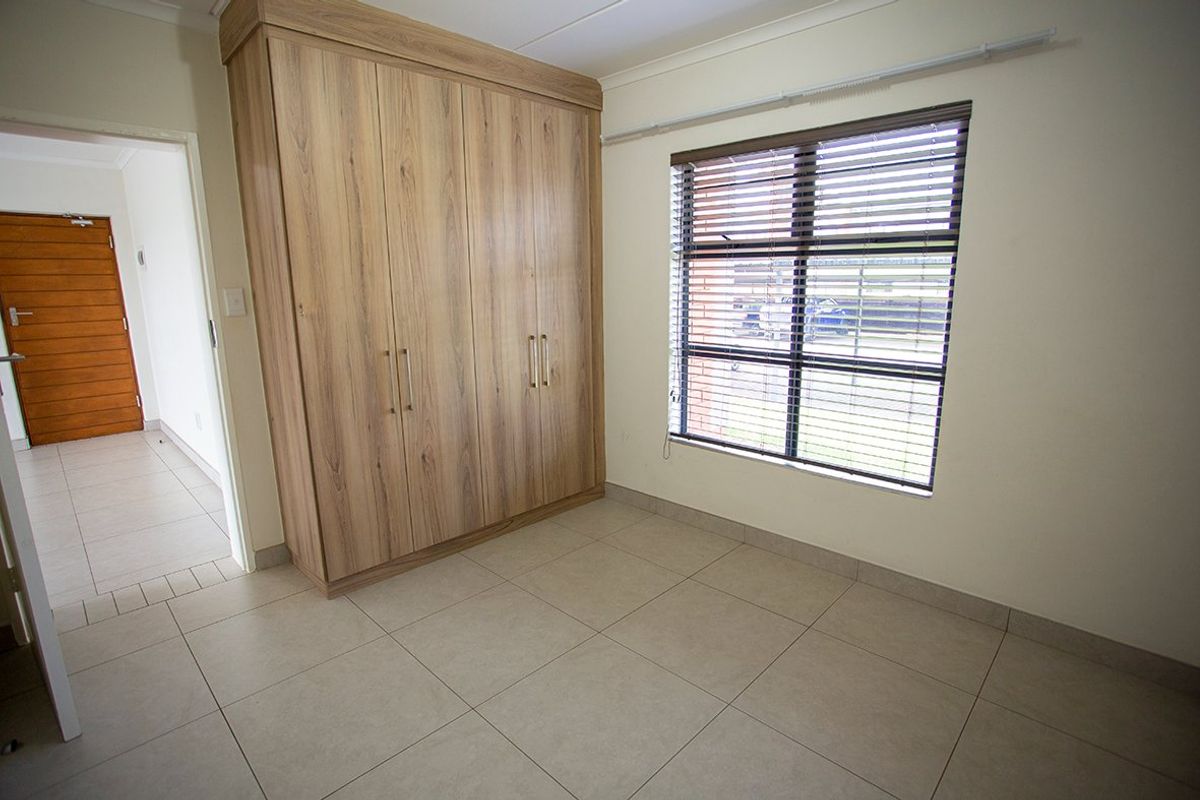 A bright, empty bedroom featuring neutral tiled flooring, a large built-in wooden wardrobe, and venetian blinds on the window.