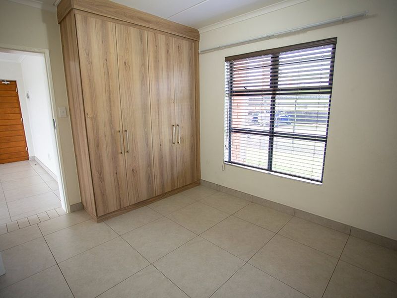 A bright, empty bedroom featuring neutral tiled flooring, a large built-in wooden wardrobe, and venetian blinds on the window.