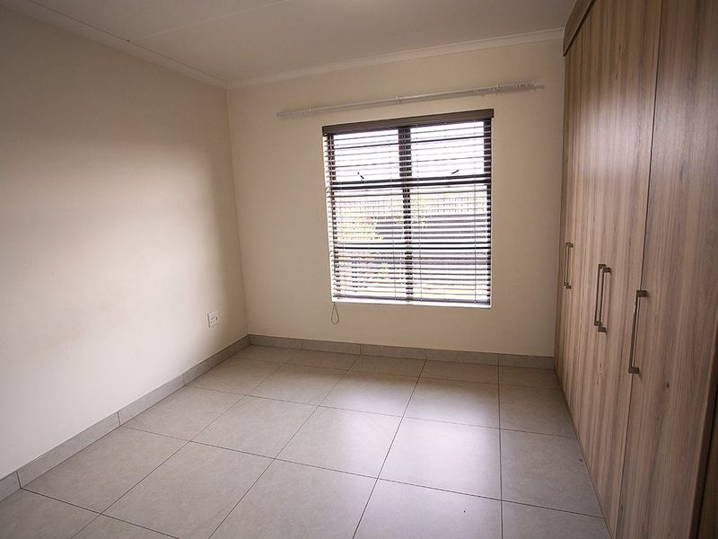 A clean, empty bedroom featuring neutral tiled flooring, built-in wooden cupboards, and venetian blinds, offering a versatile space ready for personalization.