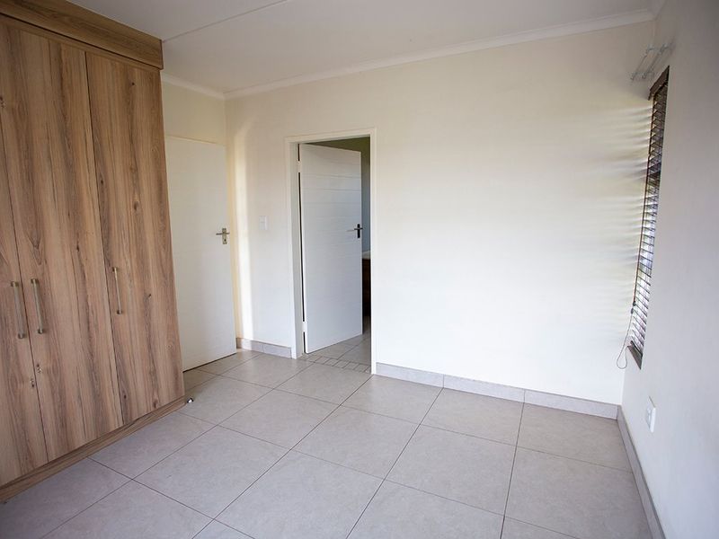 A clean, empty bedroom featuring neutral tiled flooring, a modern wood-finish built-in wardrobe, and venetian blinds.