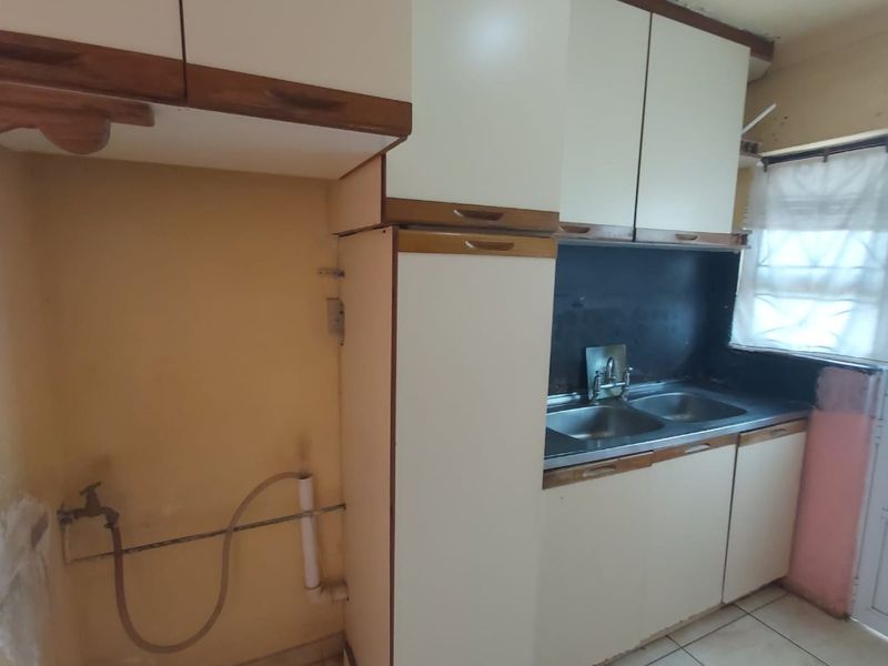 A compact kitchen area featuring built-in cabinetry, a double sink, and tiled flooring, currently requiring cosmetic updates and wall repairs.