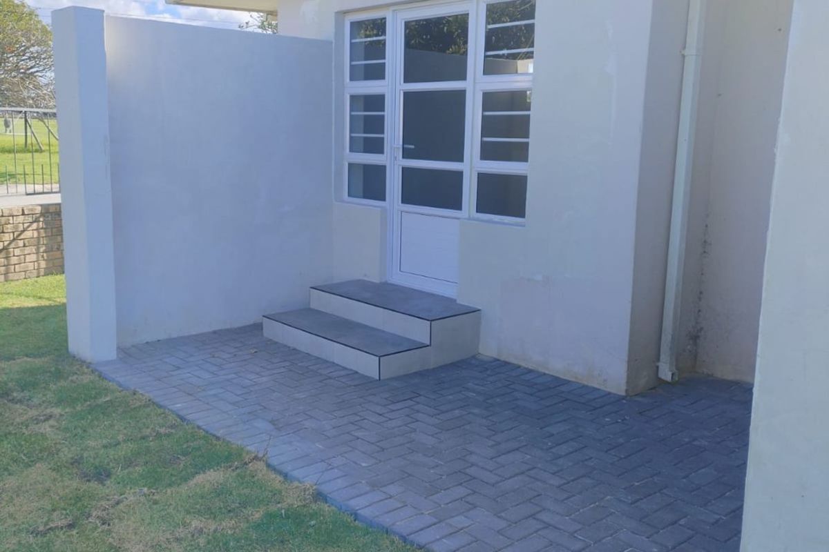 Rear exterior view of the apartment featuring a paved patio area, tiled entrance steps, and a glass panel door with external lighting.