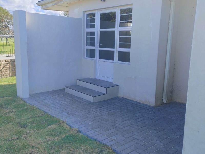 Rear exterior view of the apartment featuring a paved patio area, tiled entrance steps, and a glass panel door with external lighting.