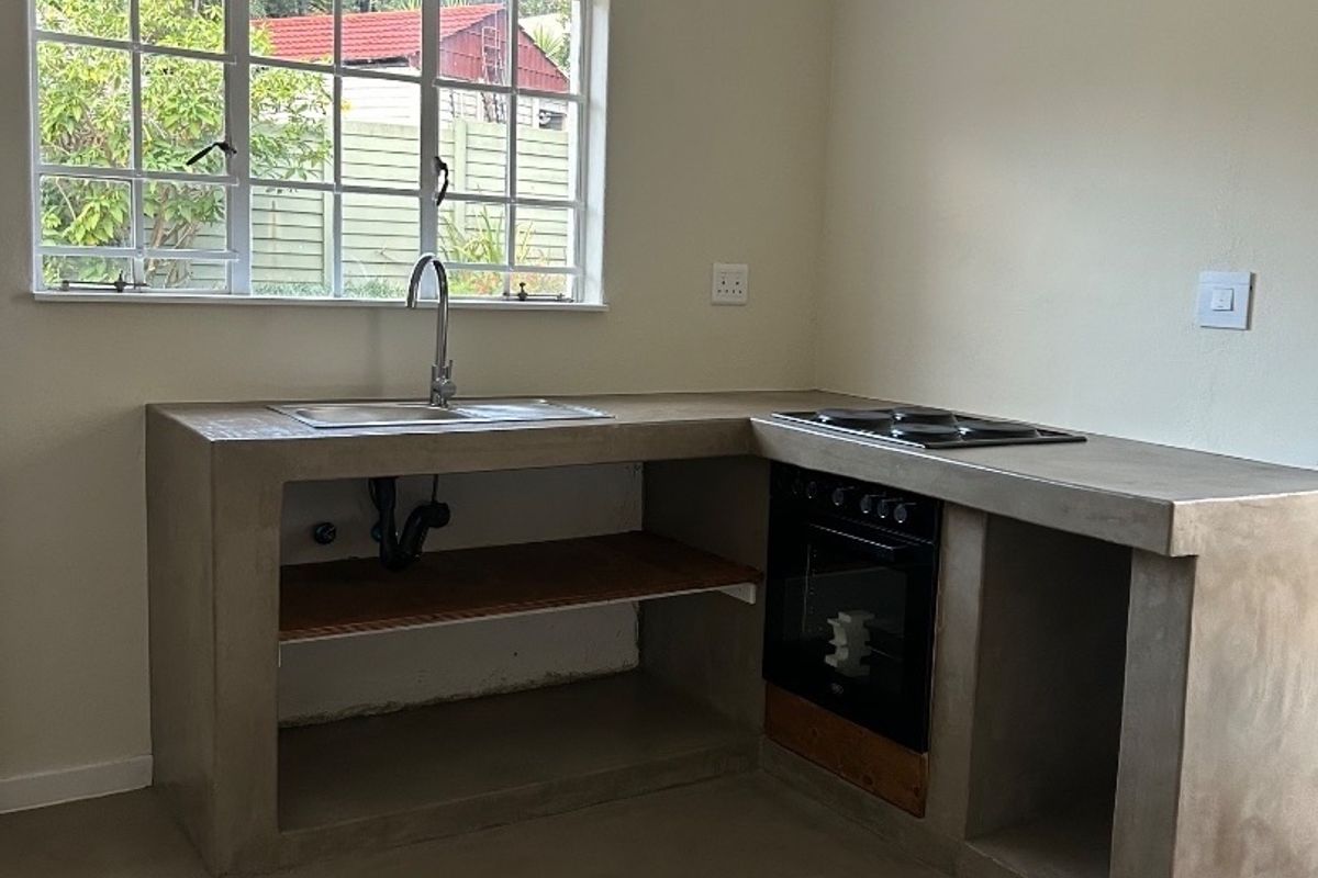 A minimalist kitchen featuring a concrete countertop, built-in oven and hob, and a stainless steel sink, currently awaiting final cabinetry finishes.