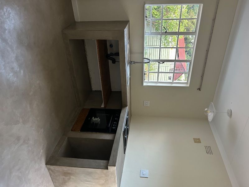 A minimalist kitchen featuring a raw concrete countertop, built-in oven, hob, and sink, set against a neutral wall with a large window.