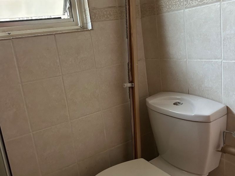 A functional bathroom featuring a standard toilet, ceramic wall tiling, and a louver window with security bars.