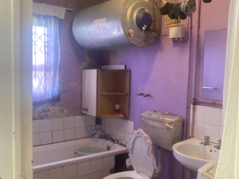 A dated bathroom featuring a bathtub, toilet, and washbasin, with visible wall surface deterioration and an exposed electric water heater.
