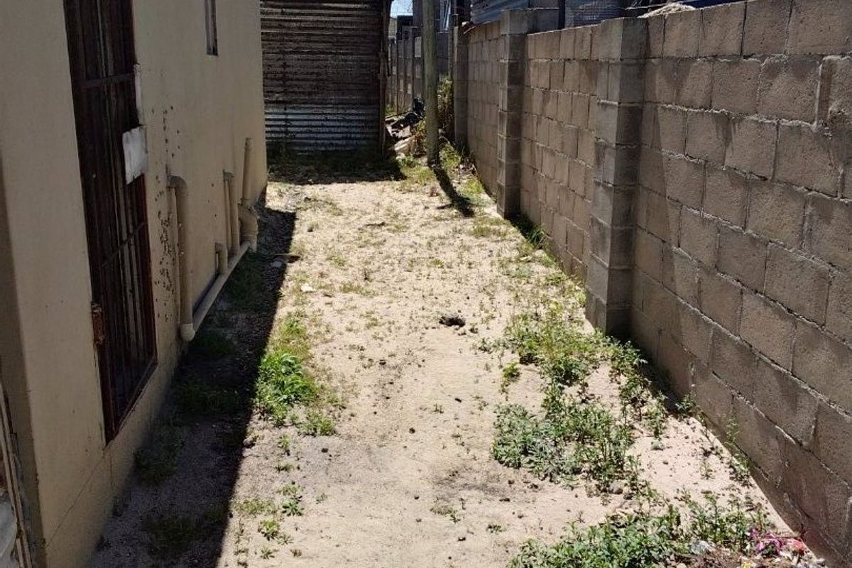 A narrow side or rear yard area featuring a concrete block boundary wall, metal security gate, and unpaved ground requiring landscaping and maintenance.