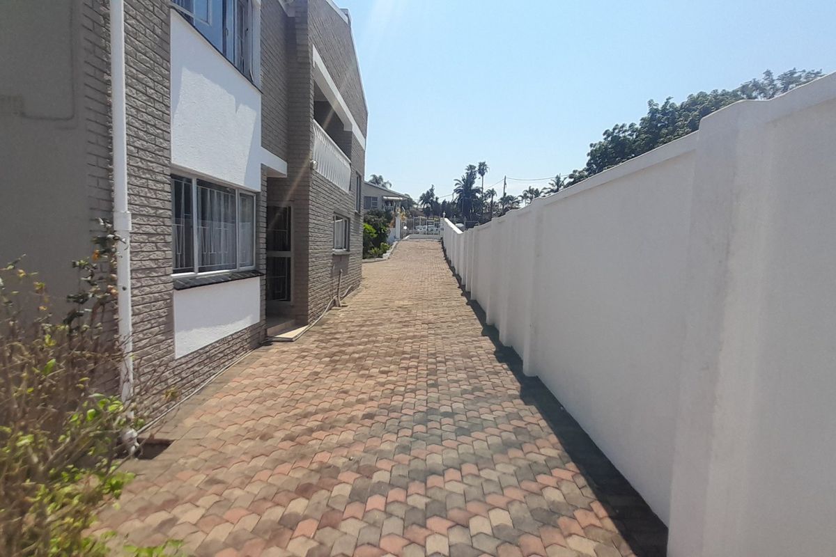 A paved side walkway leading along the side of the house, bordered by a high white boundary wall and the brick exterior of the building.