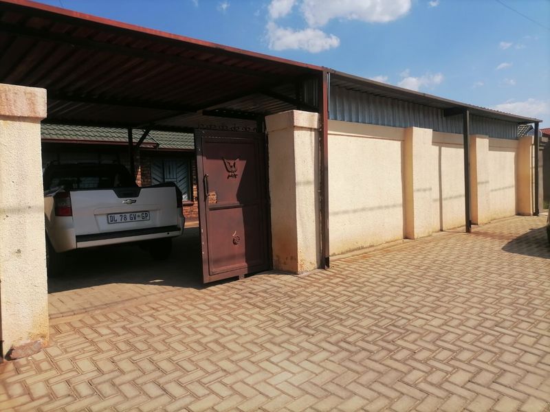 Front exterior view featuring a paved driveway, covered carport, and secure perimeter wall with a metal gate.