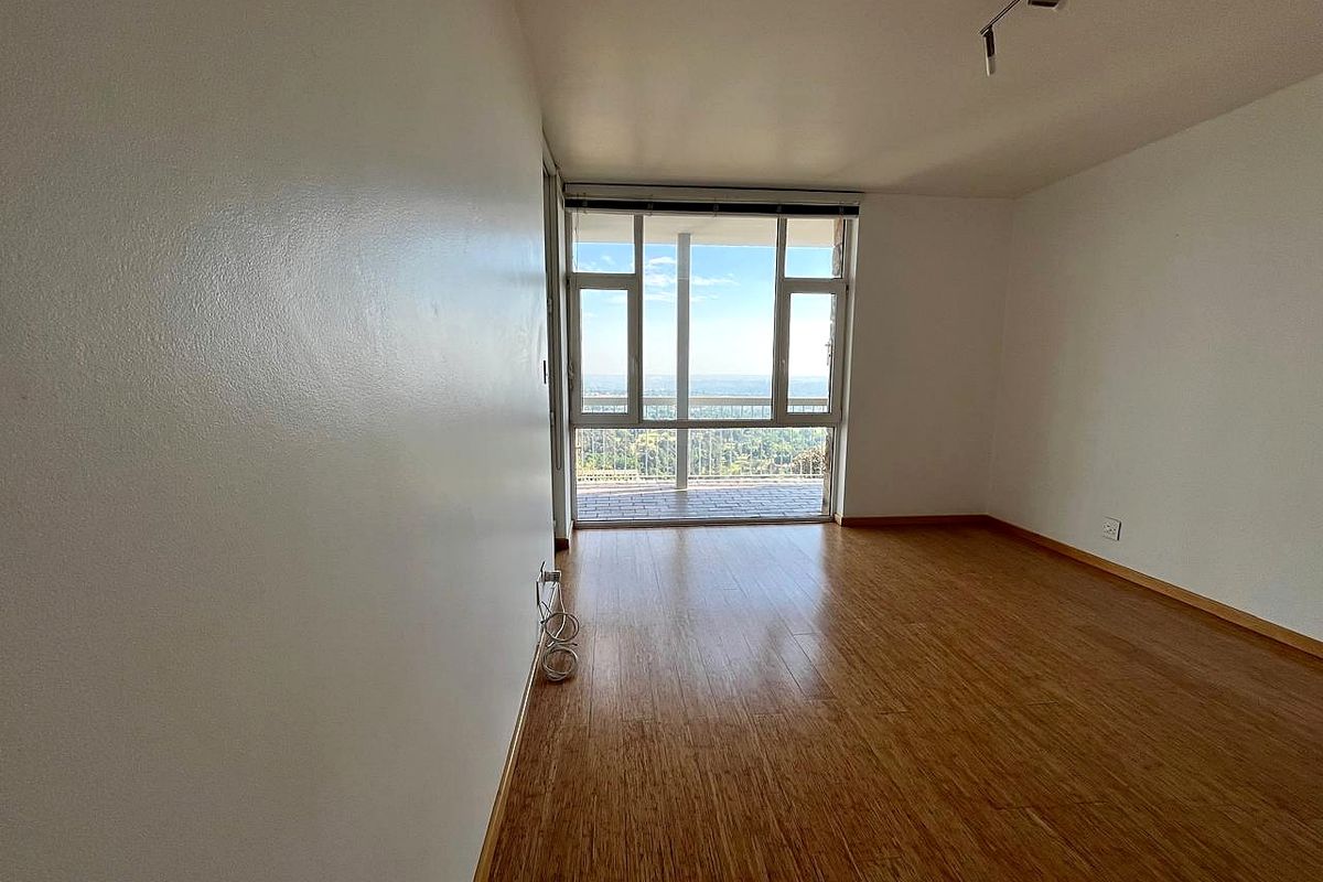 A bright, empty living room featuring laminate flooring, track lighting, and large windows providing access to a balcony with scenic views.