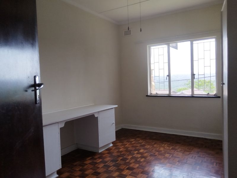 A bright, empty bedroom featuring classic parquet flooring, a built-in desk, and large windows with security bars offering a pleasant view.