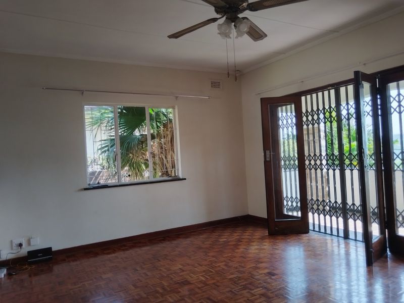 A bright, empty living room featuring classic parquet flooring, a ceiling fan, and direct access to an outdoor area through a secure timber door.