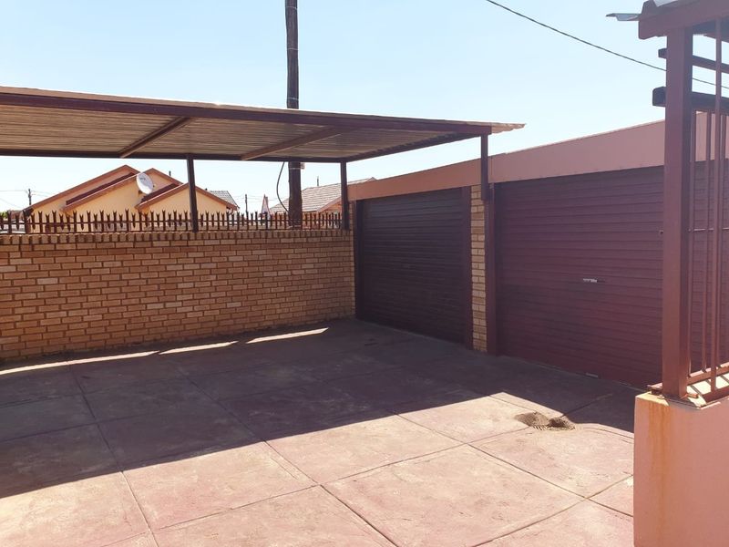 A paved driveway area featuring a covered carport and two roller shutter garage doors, enclosed by a brick boundary wall.